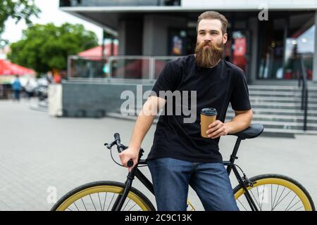 Kaffee für unterwegs. Seitenansicht des jungen bärtigen Mannes, der Kaffee trinkt, während er auf seinem Fahrrad im Freien sitzt Stockfoto