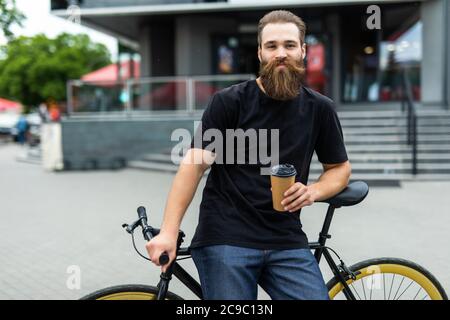 Schöner bärtiger Mann trinkt Kaffee und lächelt beim Fahrradfahren in der Stadt Stockfoto