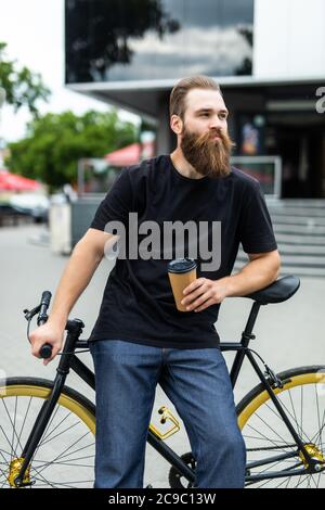 Schöner bärtiger Mann trinkt Kaffee und lächelt beim Fahrradfahren in der Stadt Stockfoto