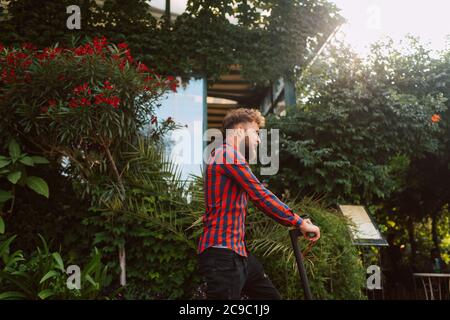 Seitenansicht eines hübschen jungen bärtigen kaukasischen Mannes mit Sonnenbrille Fahrten auf der Straße e Scooter Stockfoto