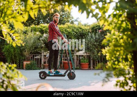 Seitenansicht eines hübschen jungen bärtigen kaukasischen Mannes mit Sonnenbrille Fahrten auf der Straße e Scooter Stockfoto