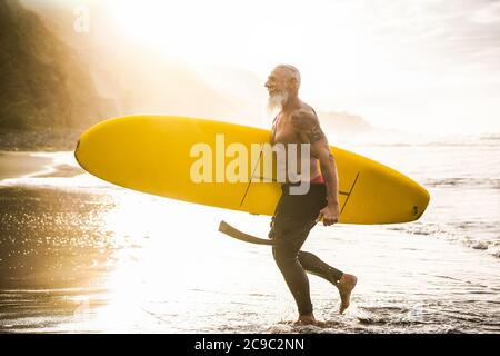 Erfahrener Fit Kerl, der nach dem Surfen bei Sonnenuntergang mit Longboard spaziert - reifer tätowierter Mann, der Spaß hat, Extremsport am tropischen Strand zu machen - fröhliche ältere Menschen Stockfoto