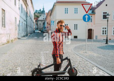 Ein hübscher junger Kaukasusmann mit Bart und Sonnenbrille geht über einen Fußgänger und hält einen Elektroroller in der Hand Stockfoto