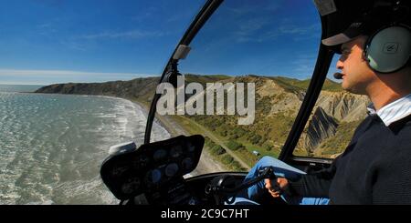 Eine weite Panoramaansicht aus der Luft in einem Robinson R44 Hubschrauber. Pilot auf der rechten Seite des Rahmens und Strand auf der linken Seite. Fliegen auf etwa 500 Fuß über einem NZ Strand. Stockfoto