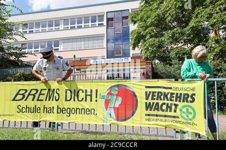 Rostock, Deutschland. Juli 2020. Matthias Bartsch, Präventionsbeauftragter der Rostocker Polizei, und Angelika Stiemer, Verkehrswache, befestigen vor der Grundschule am Mühlenteich ein Transparent mit der Aufschrift "Brems dich! Die Schule hat begonnen." Am 03.08.2020 beginnt wieder der Unterricht für rund 150,000 Schüler in Mecklenburg-Vorpommern. Quelle: Bernd Wüstneck/dpa-Zentralbild/ZB/dpa/Alamy Live News Stockfoto