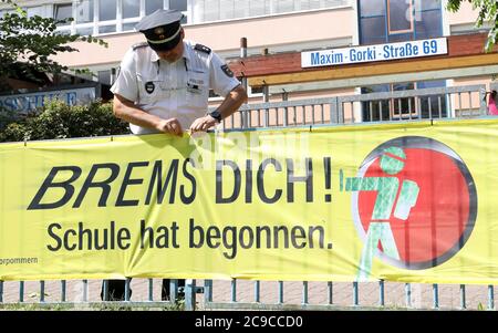 Rostock, Deutschland. Juli 2020. Matthias Bartsch, Präventionsbeauftragter der Rostocker Polizei, befestigt vor der Grundschule am Mühlenteich ein Transparent mit der Aufschrift "Brems dich! Die Schule hat begonnen." Am 03.08.2020 beginnt wieder der Unterricht für rund 150,000 Schüler in Mecklenburg-Vorpommern. Quelle: Bernd Wüstneck/dpa-Zentralbild/ZB/dpa/Alamy Live News Stockfoto