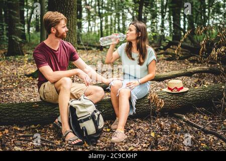 Wanderer machen Pause während Waldspaziergang. Junge kaukasische Mann und Frau sitzen auf gefallenen Baum und Trinkwasser aus Kunststoff transparente Flasche. Paar Stockfoto