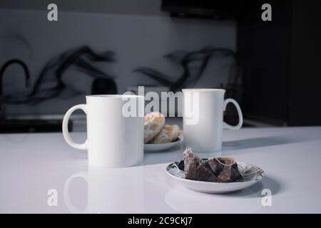 Zwei weiße Tassen Tee neben gebrauchten Teebeuteln neben Lebkuchen auf einem weißen Tisch in einer grauen Küche. Hochwertige Fotos Stockfoto