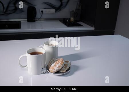 Zwei weiße Tassen mit Tee und aufragte Teebeutel neben Lebkuchen herzförmig auf einem weißen Tisch in einer grauen Küche. Hochwertige Fotos Stockfoto