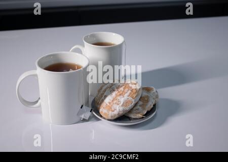 Zwei weiße Tassen mit Tee und aufragte Teebeutel neben Lebkuchen herzförmig auf einem weißen Tisch in einer grauen Küche. Hochwertige Fotos Stockfoto