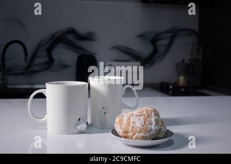 Zwei weiße Tassen mit Tee und aufragte Teebeutel neben Lebkuchen herzförmig auf einem weißen Tisch in einer grauen Küche. Hochwertige Fotos Stockfoto