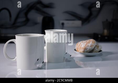 Zwei weiße Tassen mit Tee und aufragte Teebeutel neben Lebkuchen herzförmig auf einem weißen Tisch in einer grauen Küche. Hochwertige Fotos Stockfoto