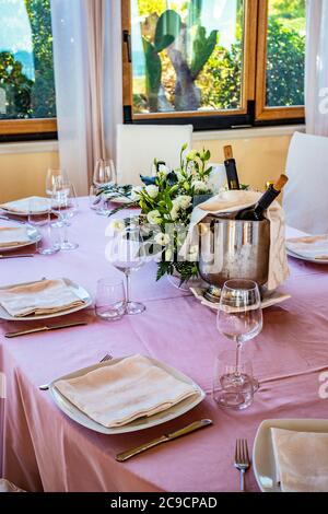 Tischset für eine Zeremonie in einem italienischen Restaurant. Feiern für eine Hochzeit. Korb mit Champagner. Blumenschmuck. Rosa Tischdecke. Luxus, Ele Stockfoto