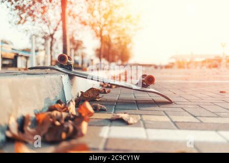 Skateboarding. Ein umgedrehtes Skateboard liegt auf dem Bürgersteig, mit heruntergefallenen Blättern. Im Hintergrund eine leere Straße der Stadt. Sonnenlicht. Speicherplatz kopieren Stockfoto