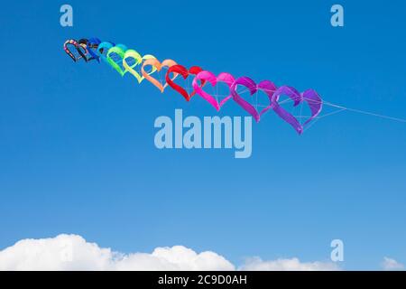 Eine Reihe von regenbogenfarbenen, herzförmigen Drachen, die am blauen Himmel über weißen Wolken fliegen. Hintergrund oder Hintergrundbild. Stockfoto