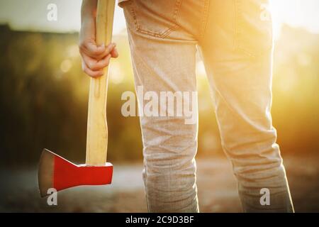 Ein Holzfäller in Jeans steht und hält in der Hand sein Werkzeug - eine Axt mit langem Griff und einer roten Klinge, die an einem warmen Sommertag von Sonnenlicht beleuchtet wird. Stockfoto