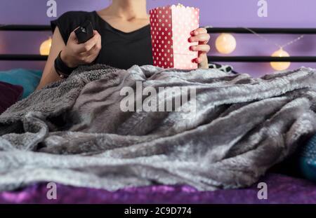 Frau im Bett mit Decke bedeckt, die Popcorn und Fernbedienung hält Stockfoto