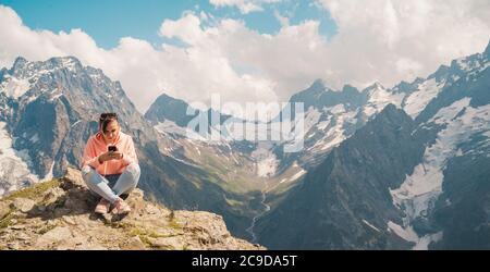 Weibliche Reisende mit Smartphone in den Bergen. Ganzkörperfrau auf Felsen sitzend und Smartphone gegen bewölkten Himmel am sonnigen Tag in den Bergen surfen Stockfoto