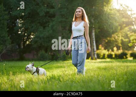 Hübsche Frau in lässigem Outfit lächelnd und auf Gras mit französisch Bulldogge an der Leine zu Fuß. Wunderschöne kaukasische Mädchen genießen Promenade mit reinrassigen Haustier im Sommer Stadtpark, Sonnenuntergang. Stockfoto