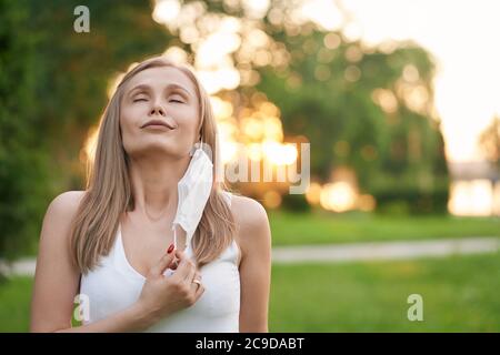 Portrait der jungen attraktiven Frau mit geschlossenen Augen, die weiße Schutzmaske auszieht und tiefes Atmen macht. Weibliches Modell steht im grünen Park, Sommeruntergang auf Hintergrund. Pandemiekonzept. Stockfoto