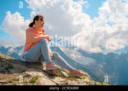 Junge Frau, die auf Felsen sitzt und die Berglandschaft anschaut. Weibliche Reisende genießen schöne Aussicht in der bergigen Gegend. Stockfoto