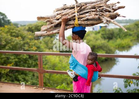 Eine Frau überquert eine Brücke über den Niger, den drittlängsten Fluss Afrikas, während sie ein Feuerholz und ihr Kleinkind auf dem Rücken in Kouroussa, Guinea, Westafrika, trägt. Stockfoto