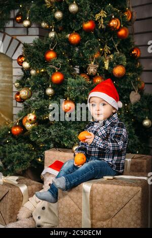 Weihnachtsporträt von glücklich lächelnd kleinen Jungen in roten weihnachtsmann Hut sitzt auf Boxen mit Geschenken halten Orangen in den Händen in der Nähe des weihnachtsbaums Stockfoto