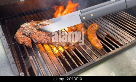 Metallzange zum Drehen von Steaks und Würstchen auf dem flammenden Gasgrill Stockfoto