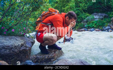 Männlicher Reisender wäscht sein Gesicht mit Bergwasser. Mann Tourist sitzt auf Felsen in der Nähe des Bergflusses und erfrischt sich. Stockfoto