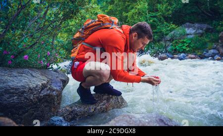 Männlicher Reisender wäscht sein Gesicht mit Bergwasser. Mann Tourist sitzt auf Felsen in der Nähe des Bergflusses und erfrischt sich. Stockfoto