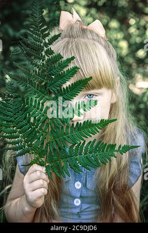 Charmantes kleines Mädchen mit blauen Augen und langen goldenen Haaren lächelt, versteckt sich hinter einem grünen Farnblatt.. Happy Funny tinn Stockfoto