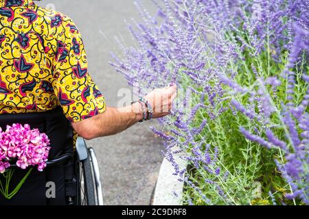 Mann im bunten Hemd sitzt in einem Rollstuhl und berührt Blumen im Freien Stockfoto