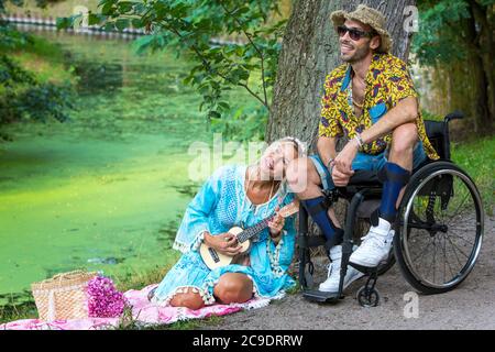 Mann im Rollstuhl und blonde Frau mit einer Gitarre, die an einem Fluss sitzt Stockfoto