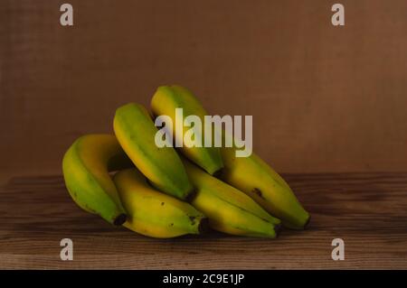 Das Stillleben der Gruppe der grünlich-gelben Bananen auf dem hölzernen Tisch, mit dem braunen Hintergrund mit dem Kopierraum Stockfoto