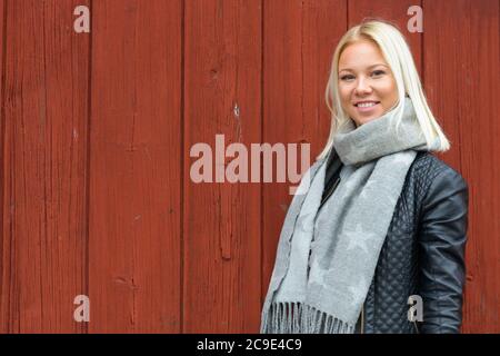 Glückliche junge schöne blonde Frau lächelt gegen rote Holzwand Stockfoto