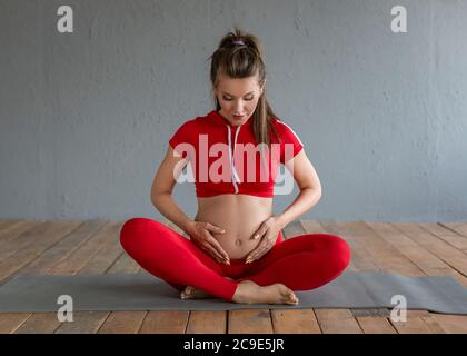 Eine schwangere Frau sitzt in Lotusposition, hält ihren Bauch mit den Händen und schaut auf ihren Bauch. Vorderansicht. Stockfoto