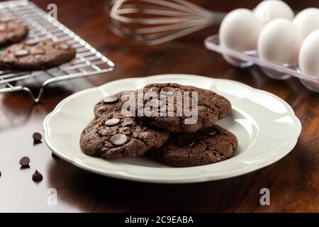 Schokoladenkakao-Kekse mit Schokoladenstückchen. Hintergrund mit Tablett und Rezept Zutaten auf Holztisch Stockfoto