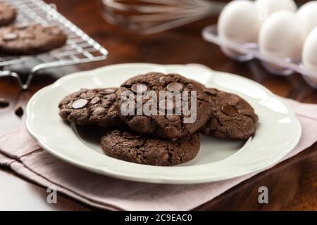 Schokoladenkakao-Kekse mit Schokoladenstückchen. Hintergrund mit Tablett und Rezept Zutaten auf Holztisch Stockfoto