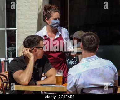 Cambridge, Großbritannien. Juli 2020. Eine Kellnerin, die eine Gesichtsmaske als vorbeugende Maßnahme trägt, dient den Kunden in einem Café im Freien. Die Menschen tragen eine Vielzahl von Gesichtsmasken im Freien in Cambridge, obwohl sie im Moment in Großbritannien nur auf öffentlichen Verkehrsmitteln und in Geschäften und öffentlichen Räumen vorgeschrieben sind. Kredit: SOPA Images Limited/Alamy Live Nachrichten Stockfoto