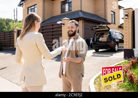 Weibliche makler in Anzug übergeben Hausschlüssel zu bärtigen Mann und gratulierte ihm mit dem Kauf Cottage House Stockfoto