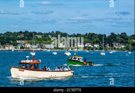 Poole, UK 06/20/2010 : EIN kleines altes grünes Fischerboot und ein Ausflugsboot segeln im Hafen mit anderen Yachten, die im Hintergrund vor Anker liegen. Als Stockfoto