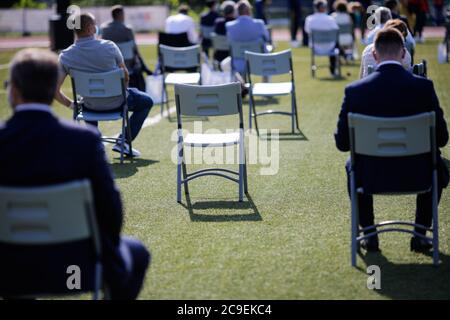 Menschen sitzen auf Stühlen auseinander, um die soziale Distanz während des Covid-19 Ausbruchs bei einem Outdoor-Event auf dem Rasen eines Stadions aufrecht zu erhalten. Stockfoto