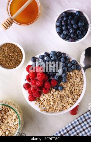 Blick von oben auf das frische Müsli-Frühstück auf dem Küchentisch. Weiße Schale mit Flocken, Heidelbeere, Himbeere und Honig auf weißer Tischdecke. Stockfoto