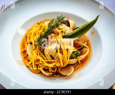 Gelbe Bandnudeln (möglicherweise "Linguine alle vongole") mit Muscheln liegen in einer leicht gewürzten Tomatensauce, garniert mit Dill und einem Salbeiblatt, auf einem tiefen weißen Teller. Im Jahr 2020, während der Coronavirus-Pandemie, wechselte das Restaurant La Cucina im Hotel Mühle in Binzen im Markgräflerland, Baden-Württemberg, Deutschland, zum ersten Mal von seiner bisherigen Markgräfler Küche zu apulischen Gerichten. Stockfoto