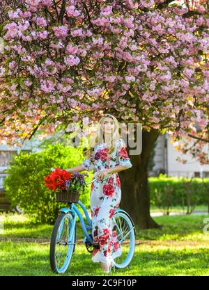 Frühlingsferien. Frau mit Tulpenstrauß. Sakura Baum blüht. Sakura-Saison. Romantische Fahrradtour. Tourismuskonzept. Transport und Reisen. Mädchen langes Kleid retro Cruiser Fahrrad Sakura Baum. Stockfoto