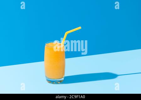Mango-Milchshake mit gelbem Stroh isoliert auf blauem Hintergrund, in hellem Licht. Ein Glas Smoothie mit Mango. Tropischer Cocktail im Sommer. Stockfoto