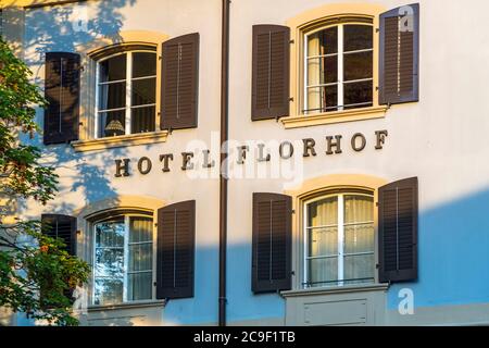 Die Schrift „HOTEL FLORHOF“ schmückt die Fassade zwischen zwei Fenstern mit dunklen Fensterläden im Abendlicht in Zürich, Schweiz. Hinter den offenen Fensterflügeln sind Vorhänge sichtbar. Warmes Licht fällt schräg über den hellblauen und beigen Putz. 1763 für die Seidenweberei-Familie Oeri-Lavater erbaut, wurde das Gebäude 1907 zur Pension Florhof. Der Hotelbetrieb endete 2022. Eine Wiedereröffnung als „Villa Florhof“ wurde angekündigt. Stockfoto