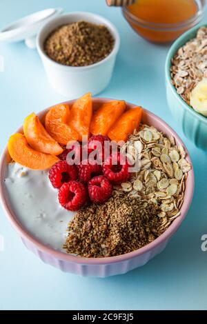 Gesundes Müsli-Frühstück auf dem blauen Tisch. Himbeere, Aprikose, Müsli, Joghurt und Leinsamen in rosa Schüssel serviert. Stockfoto