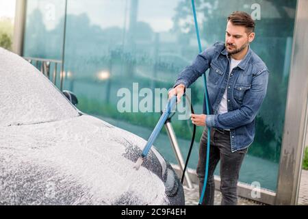 Außenreinigung des Autos mit Seifenschaum mit Hochdruckstrahl. Schöner bärtiger junger Mann, der sein Elektroauto unter hohem Druck mit Schaum wäscht Stockfoto