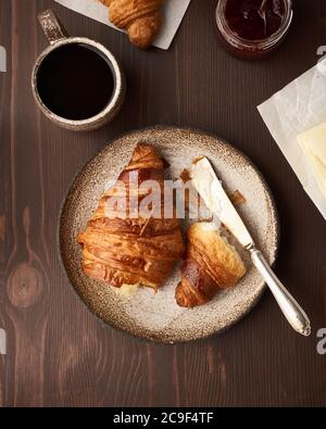 Frühstück mit Croissant und Scheibe auf dem Teller, Tasse Kaffee, Marmelade und Butter. Dunkelbrauner Tisch Stockfoto
